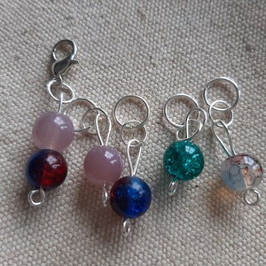 Op de afbeelding: Een verzameling handgemaakte steekmarkeerders met kleurrijke glazen kralen. De markeerders hebben roze, blauwe, rode en groene kralen en zijn bevestigd aan zilverkleurige metalen ringen en een sluiting.