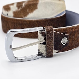May include: A brown and white cowhide belt with a silver buckle. The belt has a natural hair-on hide texture and a classic design. The buckle is rectangular and shiny. The belt is a stylish accessory.