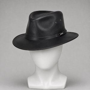 May include: A black leather fedora hat with a pinched crown and a wide brim. The hat features a matching black leather band around the base of the crown, accented with a small silver-toned metal detail and two metal eyelets on the side.