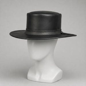 Peut inclure: Un chapeau en cuir noir doté d'un sommet plat et d'un bord large et structuré. Le chapeau présente une finition lisse et une bande en cuir noir assortie entourant la base de la calotte, fixée par un petit nœud sur le côté du bord.