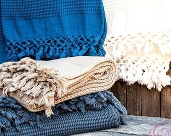 Può includere: Una pila di tre asciugamani in tessuto a nido d'ape blu, beige e grigio con dettagli a frangia. Gli asciugamani sono piegati e disposti su una superficie in legno.