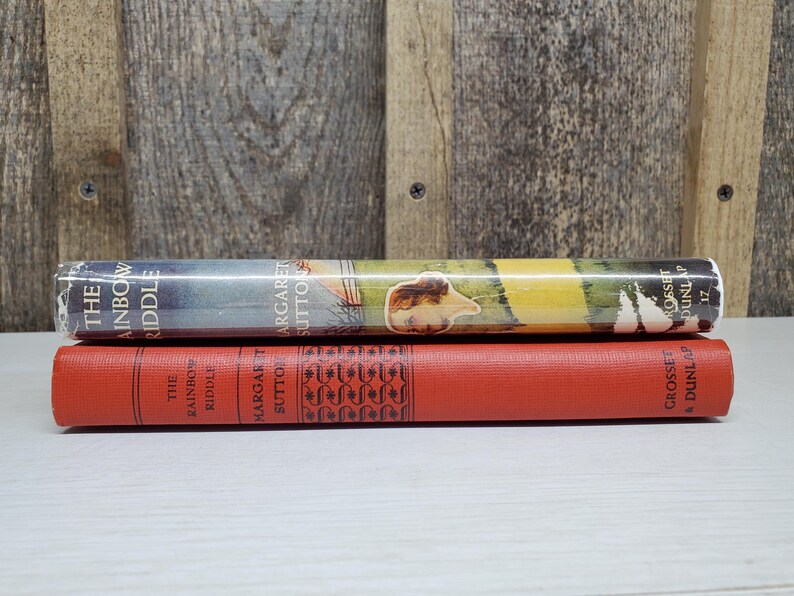 May include: Two vintage books are stacked on a white surface. The top book, "The Rainbow Riddle," has a colorful cover with a landscape scene. The bottom book is red with the title "The Rainbow Riddle" and decorative patterns.