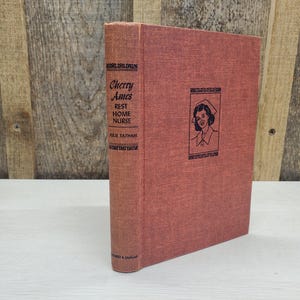 May include: A vintage book titled "Cherry Ames, Rest Home Nurse" by Julie Tatham. The book has a reddish-orange cover with a drawing of a nurse. The book is standing upright against a wooden background.