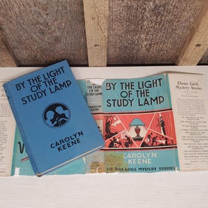 May include: A collection of vintage books, including "By the Light of the Study Lamp" by Carolyn Keene. The books are in various shades of blue and white, with text and illustrations on the covers. The books are part of the Dana Girls Mystery Stories series.