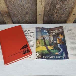 May include: A collection of vintage books, including a red hardcover titled "A Judy Bolton Mystery" and a book titled "The Rainbow Riddle" by Margaret Sutton. The covers feature illustrations and text.