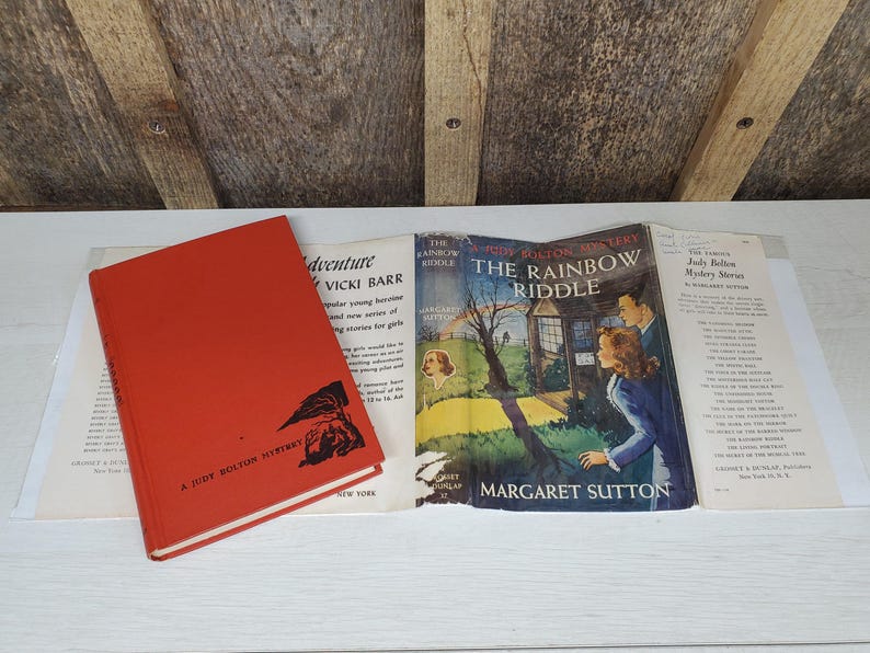 May include: A collection of vintage books, including a red hardcover titled "A Judy Bolton Mystery" and a book titled "The Rainbow Riddle" by Margaret Sutton. The covers feature illustrations and text.