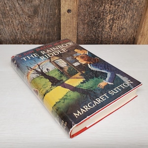 May include: A hardcover book titled "The Rainbow Riddle" by Margaret Sutton. The book cover features a colorful illustration of a house, a couple, and a tree. The book has a red spine and white pages.