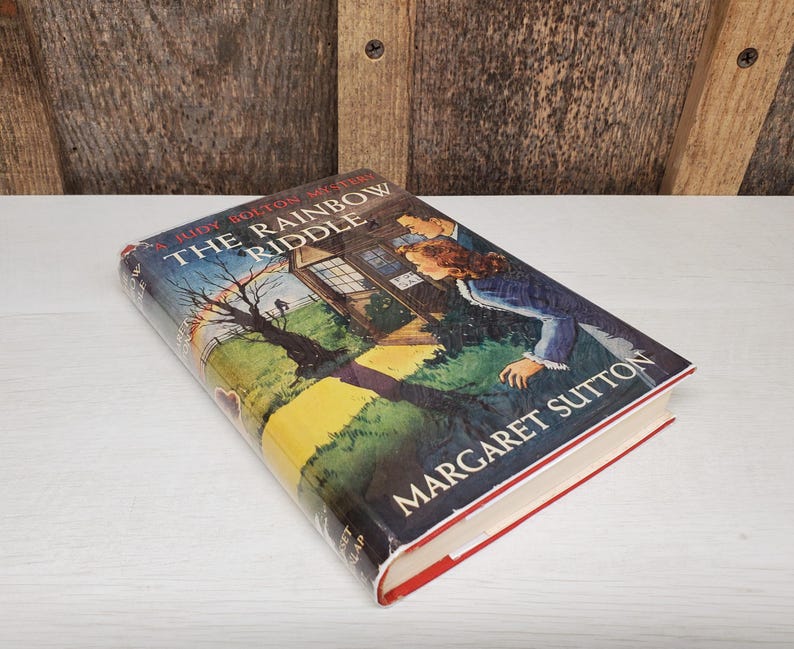 May include: A hardcover book titled "The Rainbow Riddle" by Margaret Sutton. The book cover features a colorful illustration of a house, a couple, and a tree. The book has a red spine and white pages.