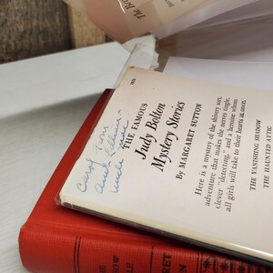 May include: Two vintage books are stacked, with the top book titled "Judy Bolton Mystery Stories" by Margaret Sutton. The cover is white with blue text and handwriting. The bottom book is red.