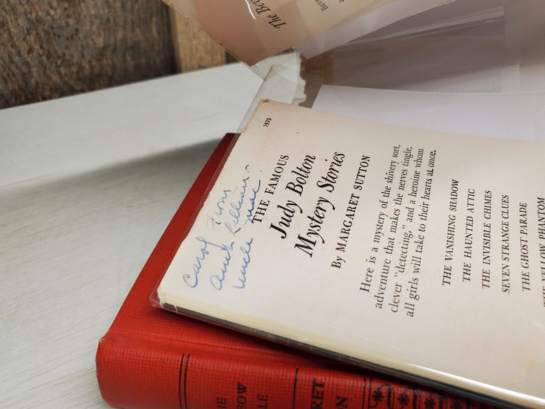 May include: Two vintage books are stacked, with the top book titled "Judy Bolton Mystery Stories" by Margaret Sutton. The cover is white with blue text and handwriting. The bottom book is red.