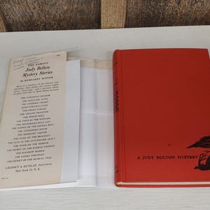 May include: A vintage red hardcover book titled "A Judy Bolton Mystery" with a black illustration. Beside it, a paper with the title "Judy Bolton Mystery Stories" and a list of titles.