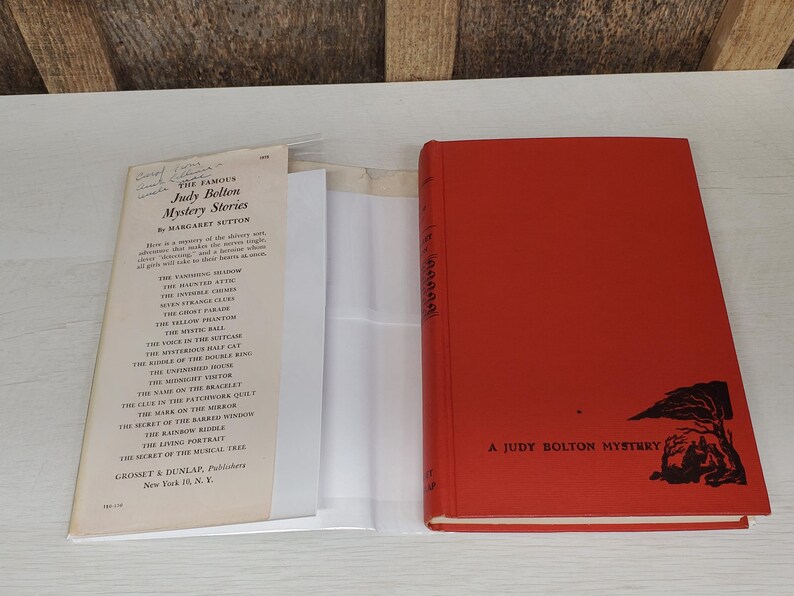 May include: A vintage red hardcover book titled "A Judy Bolton Mystery" with a black illustration. Beside it, a paper with the title "Judy Bolton Mystery Stories" and a list of titles.