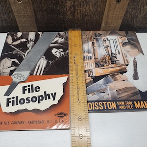May include: Vintage "File Filosophy" and "Disston Saw, Tool and File Manual" books. The "File Filosophy" book has an orange and white cover with a black and white image. The "Disston" manual has a blue cover with a black and white image. A wooden ruler is next to the books.