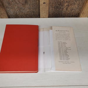 May include: An open book with a bright red cover, next to a printed page titled "The Beverly Gray Stories". The book's pages are visible, along with a clear plastic sheet. The background is a light-colored wooden surface.