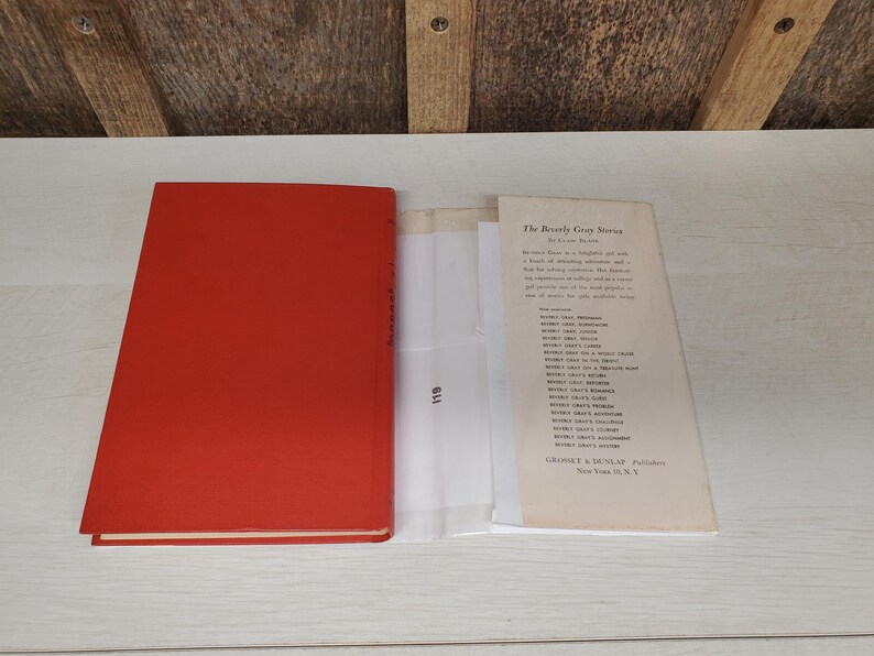 May include: An open book with a bright red cover, next to a printed page titled "The Beverly Gray Stories". The book's pages are visible, along with a clear plastic sheet. The background is a light-colored wooden surface.