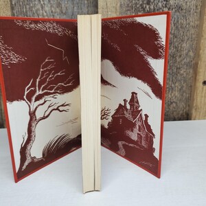May include: Open vintage book with a red cover and illustrations. One side features a bare tree and lightning, the other a spooky house. The pages are cream-colored, and the edges are red.