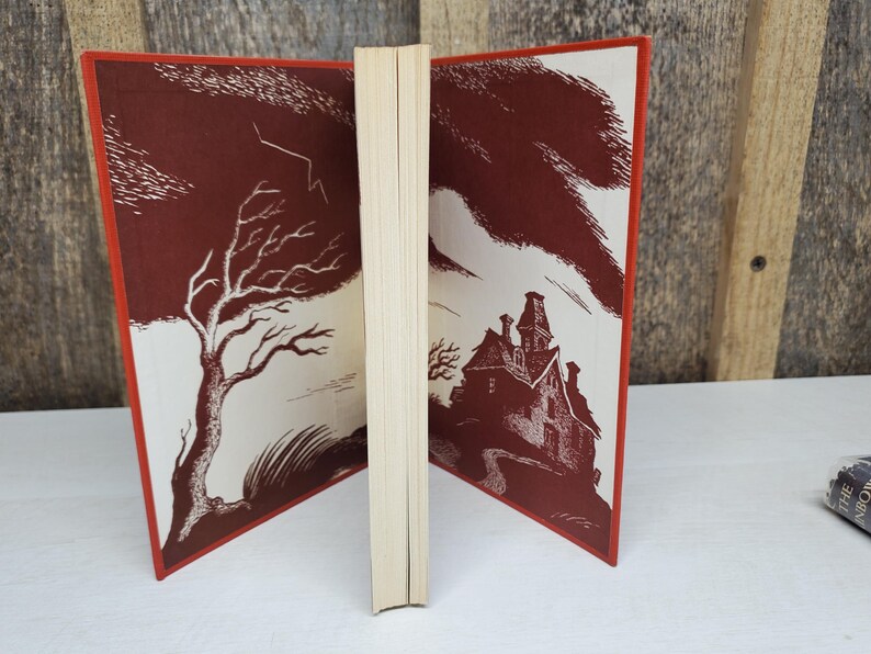 May include: Open vintage book with a red cover and illustrations. One side features a bare tree and lightning, the other a spooky house. The pages are cream-colored, and the edges are red.