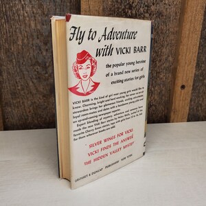 May include: A vintage book titled "Fly to Adventure with Vicki Barr." The cover features a red and white illustration of a woman in a hat. The book is a part of a series of exciting stories for girls.