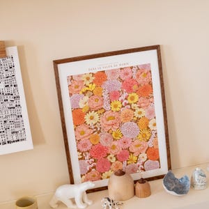 Peut inclure: Une affiche encadrée avec un cadre en bois marron, présentant un arrangement floral vibrant de fleurs roses, oranges et jaunes. L'affiche est intitulée "DANS LE SALON DE MAMIE". Des objets décoratifs, dont une figurine d'ours polaire blanc, sont exposés.