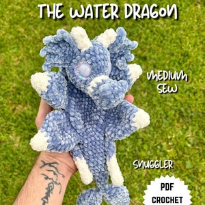 Könnte beinhalten: Ein blau-weißes Plüschdrachenspielzeug mit dem Text "ECHO THE WATER DRAGON". Der Drache hat ein großes, funkelndes Auge und ist mit "Snuggler" und "PDF Crochet Pattern" beschriftet. Das Spielzeug wird vor einem grünen Hintergrund gehalten.