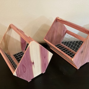 May include: Two wooden storage baskets with a handle. The baskets are made of cedar wood with a natural reddish-brown color and a wire mesh interior. The baskets are angled and have a handle for carrying.