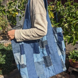 May include: A large denim tote bag with a patchwork design. The bag is made from various shades of blue denim, creating a textured and visually interesting pattern. The bag has long straps for carrying over the shoulder.