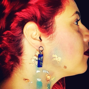 Puede incluir: Una mujer con el pelo rojo lleva un par de pendientes azules y plateados. Los pendientes son rectangulares y tienen una superficie texturizada. La cara de la mujer está parcialmente cubierta de pintura dorada y azul.