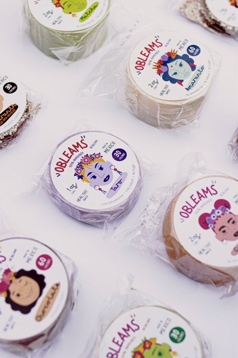 Op de afbeelding: Assortiment van individueel verpakte pakketten met "OBLEAMS" dunne, knapperige wafels. Elk pakket heeft een kleurrijke illustratie van een vrouw met een bloemenkrans en de smaaknaam. De smaken omvatten Matcha, Amaranto, Taro en Chocolade.