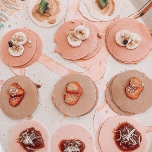 Op de afbeelding: Een assortiment mini-pannenkoeken met verschillende toppings, waaronder aardbeien, kiwi, banaan en chocolade. De pannenkoeken zijn gerangschikt op een wit oppervlak met roze en bruine accenten.