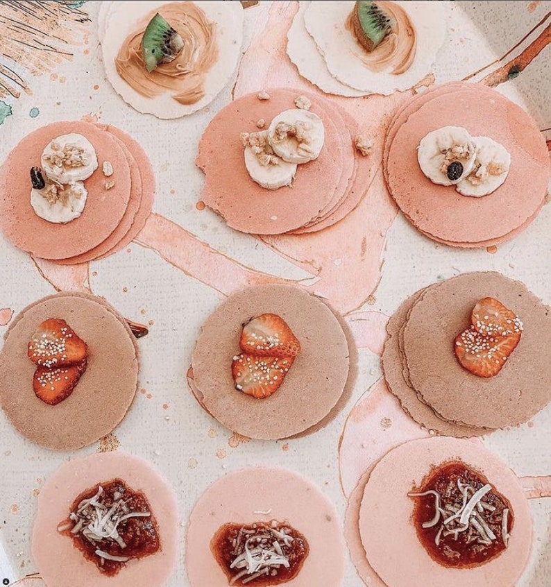 Op de afbeelding: Een assortiment mini-pannenkoeken met verschillende toppings, waaronder aardbeien, kiwi, banaan en chocolade. De pannenkoeken zijn gerangschikt op een wit oppervlak met roze en bruine accenten.