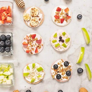 Op de afbeelding: Zes mini rijstkoekjes bedekt met yoghurt, granola, fruit en noten. De rijstkoekjes zijn gerangschikt in twee rijen van drie op een wit marmeren oppervlak. De toppings omvatten aardbeien, blauwe bessen, bananen, appelschijfjes en granola.