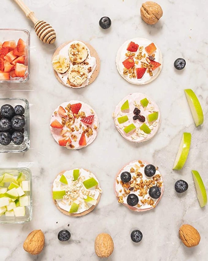 Op de afbeelding: Zes mini rijstkoekjes bedekt met yoghurt, granola, fruit en noten. De rijstkoekjes zijn gerangschikt in twee rijen van drie op een wit marmeren oppervlak. De toppings omvatten aardbeien, blauwe bessen, bananen, appelschijfjes en granola.