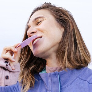 Op de afbeelding: Een vrouw met lang bruin haar glimlacht terwijl ze een paarse tongschraper voor haar mond houdt.