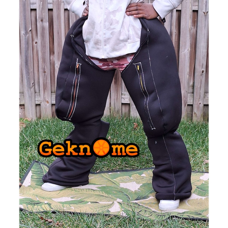 May include: Black pants with a zipper detail on each leg. The pants are made of a thick, stretchy material and have a relaxed fit. The pants are worn by a person standing on a patterned rug.