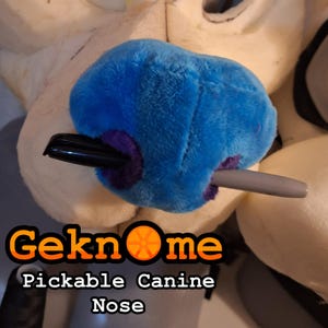May include: A blue plush canine nose with purple stitching. The nose is being held in place with a black pen and a gray pen. The text "Geknome Pickable Canine Nose" is visible below the nose.
