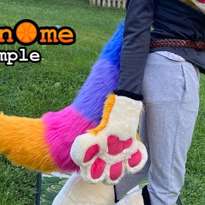 May include: A colorful furry tail and paw costume with orange, pink, purple, and blue sections. The paw has pink pads and is cream-colored. The image includes the text "Geknome Sample" in an orange and black font.