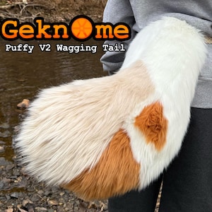 Op de afbeelding: Een pluizige, veelkleurige staart met witte, bruine en bruine vacht. De staart is voorzien van de tekst "Geknome Puffy V2 Wagging Tail" in oranje en zwarte letters. De staart wordt vastgehouden door een persoon die een grijze trui draagt.