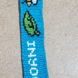 May include: A blue friendship bracelet with white lettering that says "I know" and a bee and leaf design. The bracelet has a black button with a white cross on it.