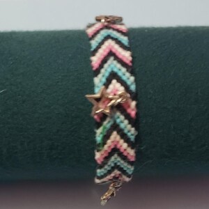May include: A black, white, pink and blue friendship bracelet with a chevron pattern and two silver star charms.