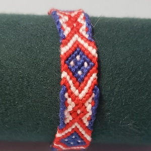 May include: Handmade friendship bracelet featuring a geometric pattern in red, white, and blue. The woven design includes diamond shapes and zigzags, with blue edges. This accessory is perfect for everyday wear.