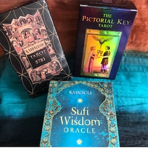 May include: Three different tarot and oracle card decks. The first deck is black with gold accents and the text "Kabbalistic Tarot 5781". The second deck is blue with rainbow accents and the text "The Pictorial Key Tarot". The third deck is blue with gold accents and the text "Rassouli Sufi Wisdom Oracle".