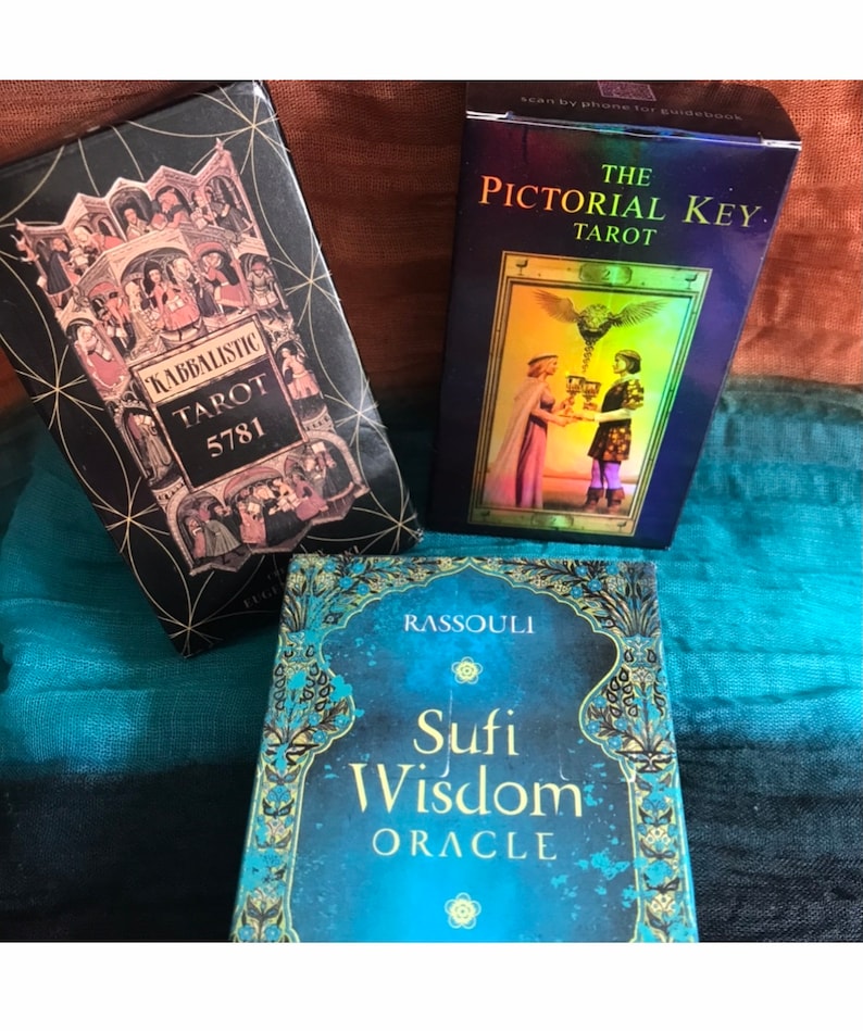 May include: Three different tarot and oracle card decks. The first deck is black with gold accents and the text "Kabbalistic Tarot 5781". The second deck is blue with rainbow accents and the text "The Pictorial Key Tarot". The third deck is blue with gold accents and the text "Rassouli Sufi Wisdom Oracle".