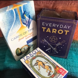 May include: Three tarot card decks, one with a blue background and the words "CARDS OF TIME Find the right moment" and a pocket watch, one with a blue background and the words "EVERYDAY TAROT" and a pentagram, and one with a white background and a woman holding two wands in a circle of green leaves.
