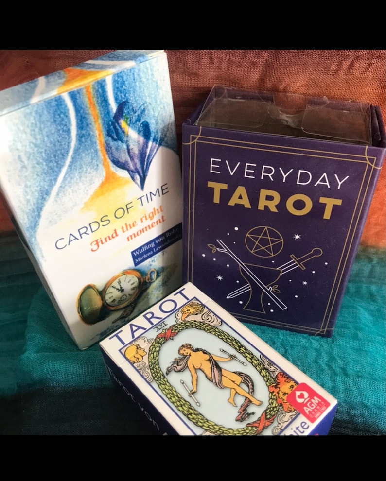 May include: Three tarot card decks, one with a blue background and the words "CARDS OF TIME Find the right moment" and a pocket watch, one with a blue background and the words "EVERYDAY TAROT" and a pentagram, and one with a white background and a woman holding two wands in a circle of green leaves.
