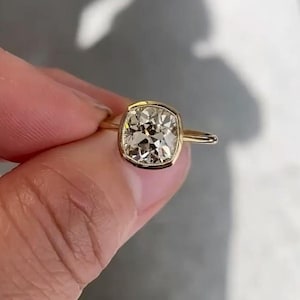 May include: A gold ring featuring a large, square-cut diamond with a light champagne hue. The diamond is set in a bezel setting, and the band is a thin, polished gold. The ring is held between fingers.