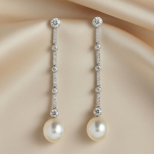 May include: A pair of elegant drop earrings. Each earring features a large, lustrous, round pearl at the bottom, topped by a series of sparkling, round, clear gemstones. The gemstones are connected by a delicate, silver-toned chain, with a larger gemstone at the top.