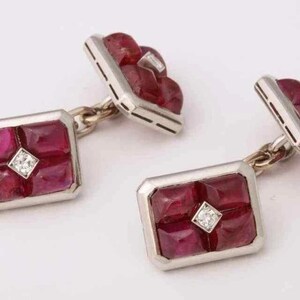 May include: A pair of rectangular cufflinks with a silver-colored frame. Each cufflink features a square arrangement of deep red gemstones, with a small diamond at the center. The cufflinks are connected by a delicate chain.