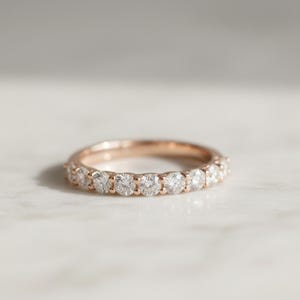 May include: Rose gold band ring with a row of round, clear diamonds. The ring is set on a white marble surface. The diamonds sparkle, reflecting light. The ring is a classic design, suitable for various occasions.
