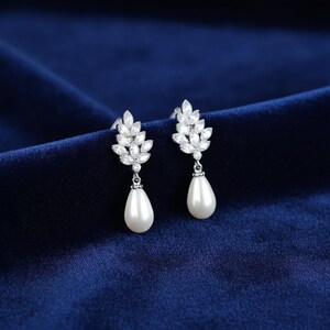 May include: A pair of silver-toned drop earrings. Each earring features a leaf-like design encrusted with small, clear crystals and a pear-shaped, white pearl drop. The earrings are set against a dark blue velvet background.