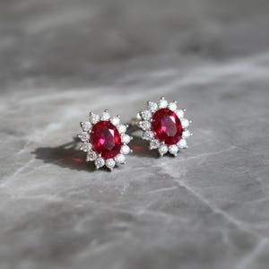May include: A pair of stud earrings featuring oval-cut, deep red gemstones surrounded by a halo of smaller, clear stones. The earrings are set in a silver-colored metal and rest on a gray surface.